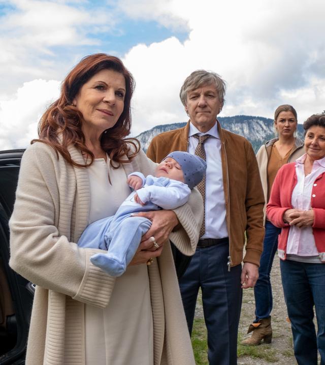 "Der Bergdoktor: Extreme": Vor dem Gruberhof: Thea Hochstetter (Claudia Wenzel) hält ein Baby auf dem Arm und lächelt. Im Hintergrund stehen Dr. Herold (Lutz Blochberger), Anne (Ines Lutz), Lisbeth (Monika Baumgartner), Lilli (Ronja Forcher) und Hans (Heiko Ruprecht) und blicken zu den beiden.