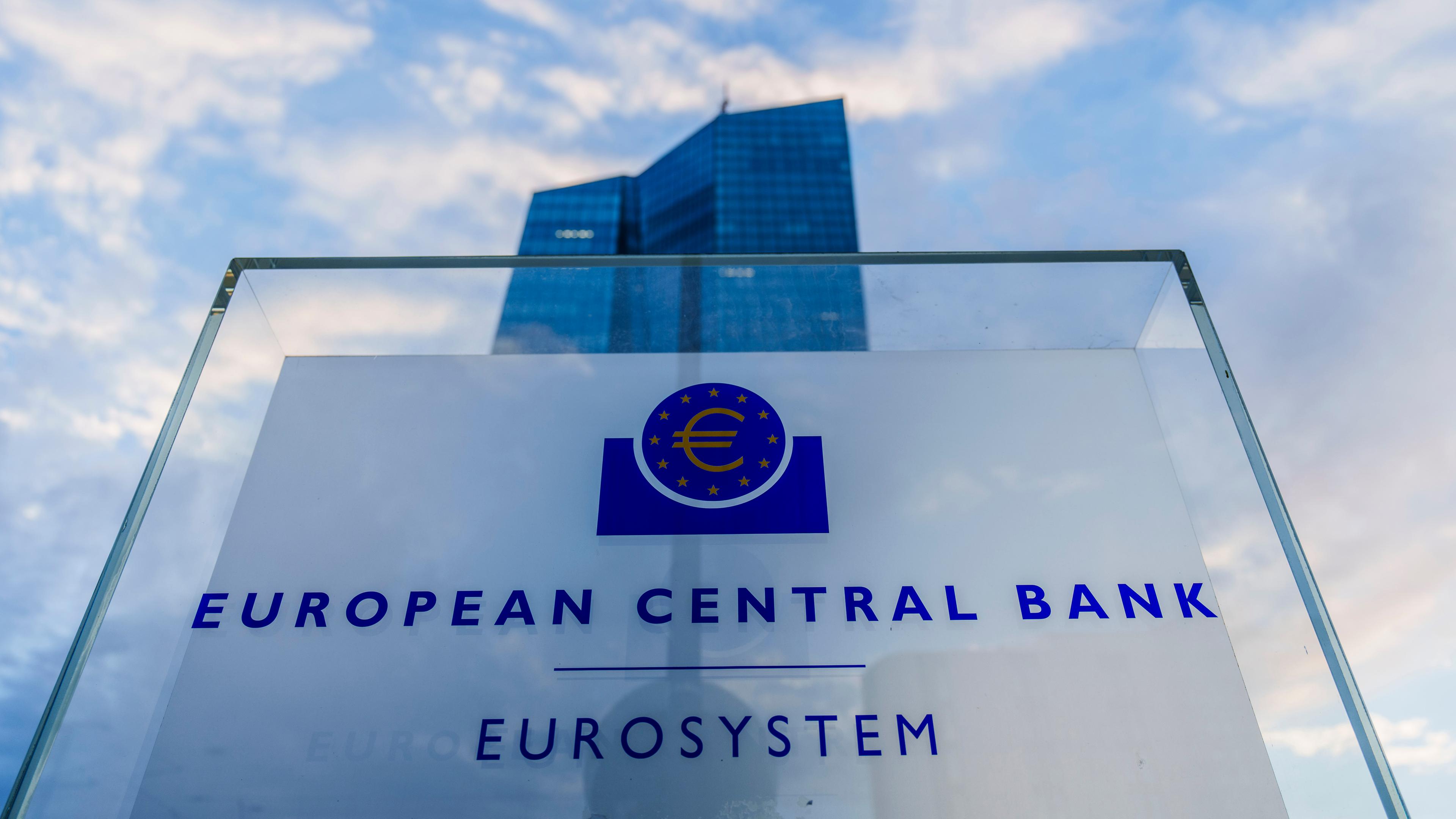 Ein Schild mit der Aufschrift "European Central Bank - Eurosystem". Im Hintergrund die Zentrale der EZB.