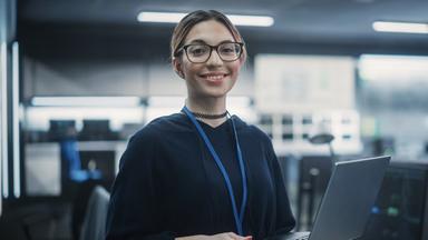 Typical für eine Aubilsung als Fachinformatiker Sytemintegration im ZDF: Junger Frau lächelnd mit einem Laaptopn in der Hand in einem Raum mit Servern