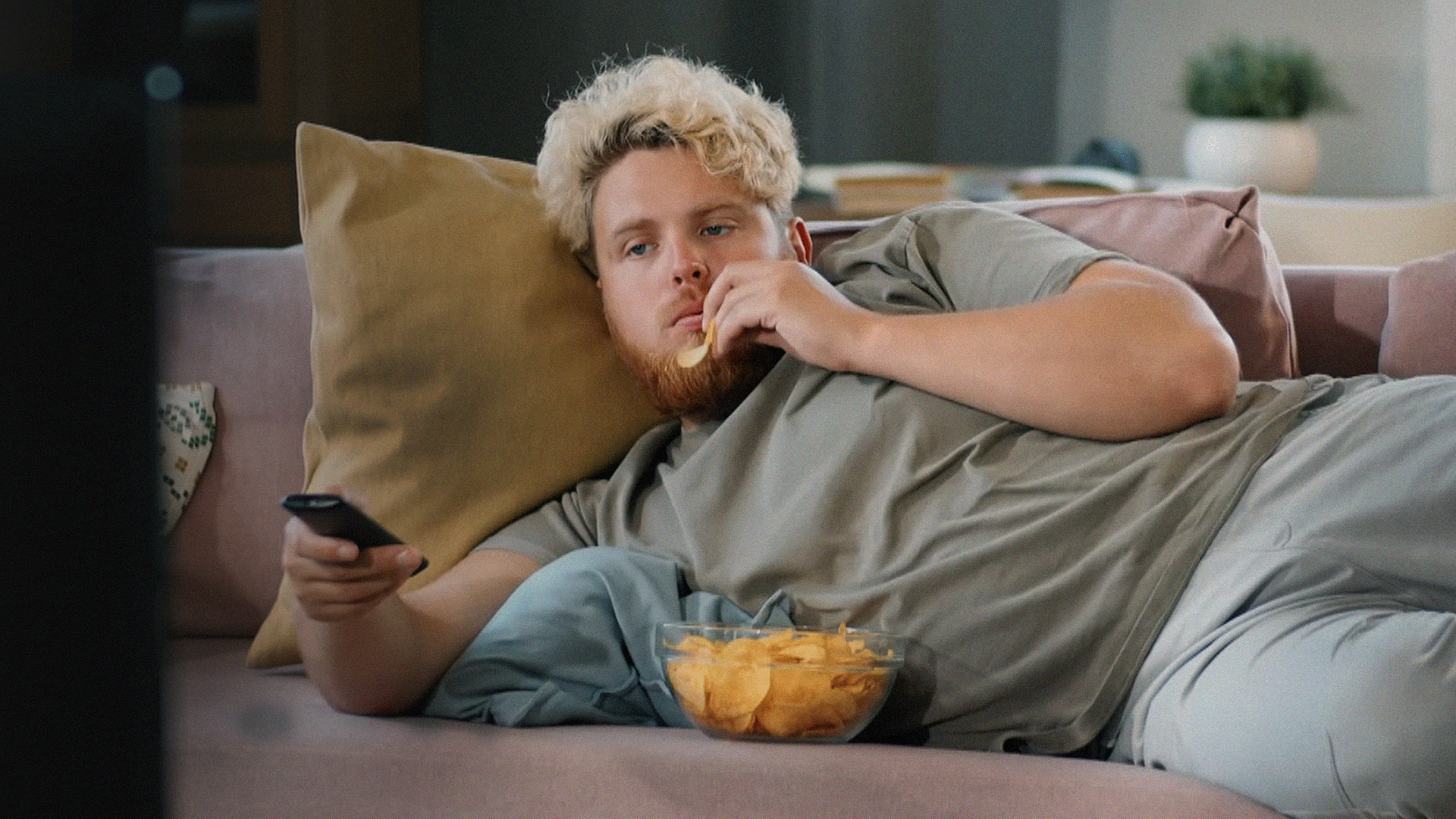 Junger Mann (Gen Z) liegt auf der Couch und isst Chips