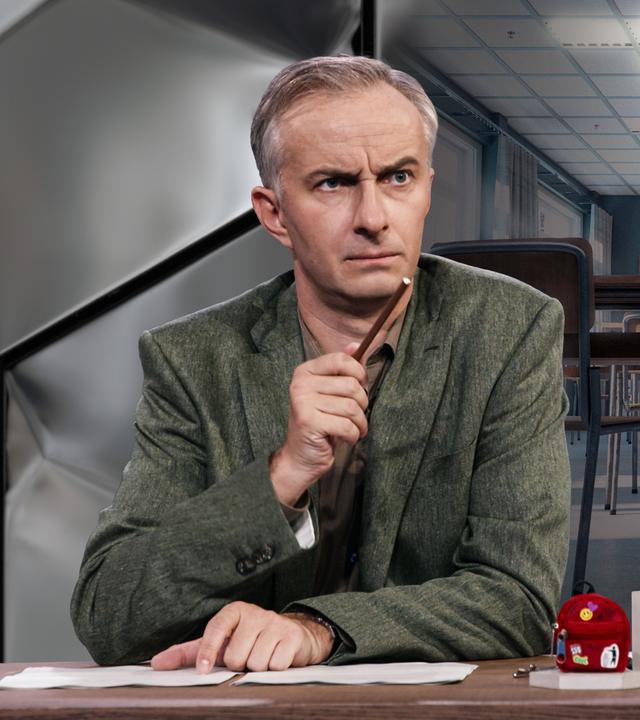 Jan Böhmermann sitzt am Schreibtisch. Im Hintergrund ist ein Bild eines leeren Klassenzimmers zu sehen.