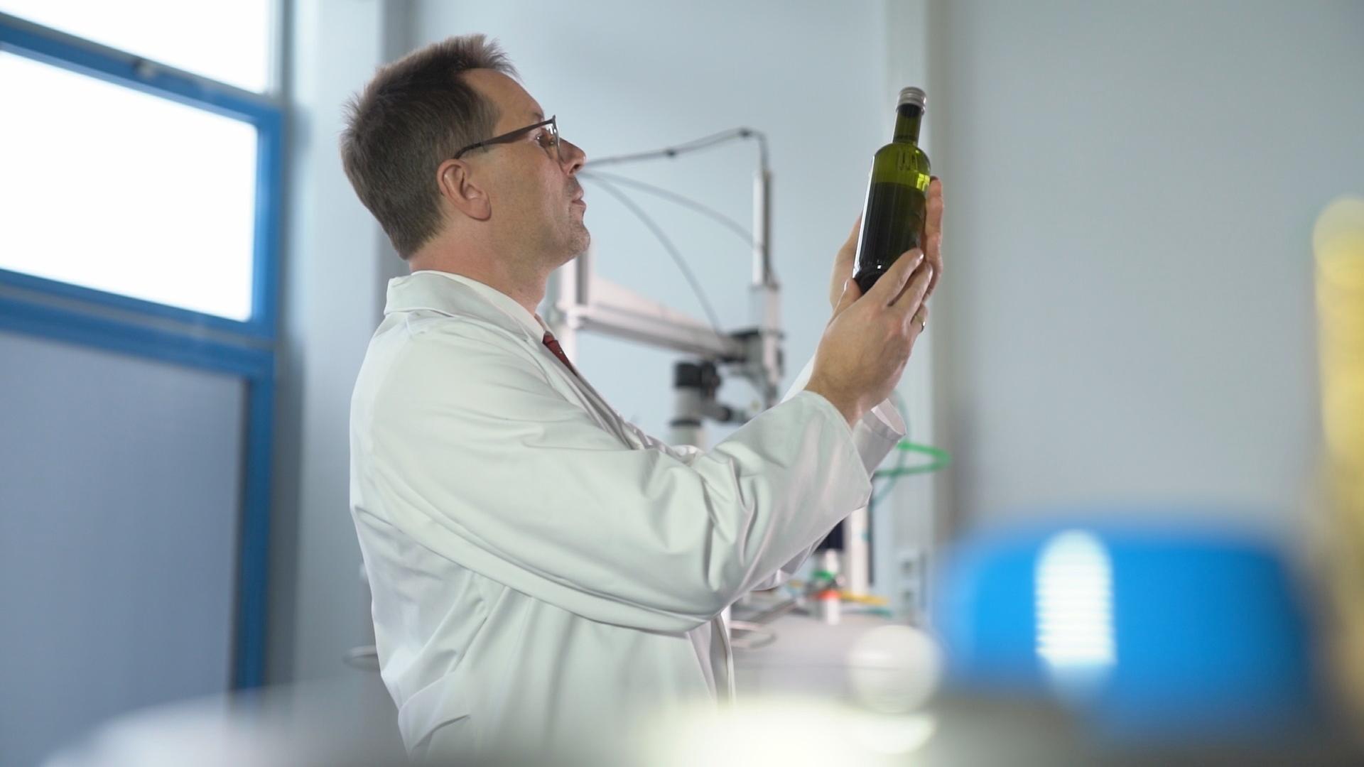 "Fake Food - Die Tricks der Lebensmittelfälscher": Prof. Stephan Schwarzinger von der Universität Bayreuth steht in einem Labor und betrachtet eine Flasche Olivenöl, die er in der Hand hält.