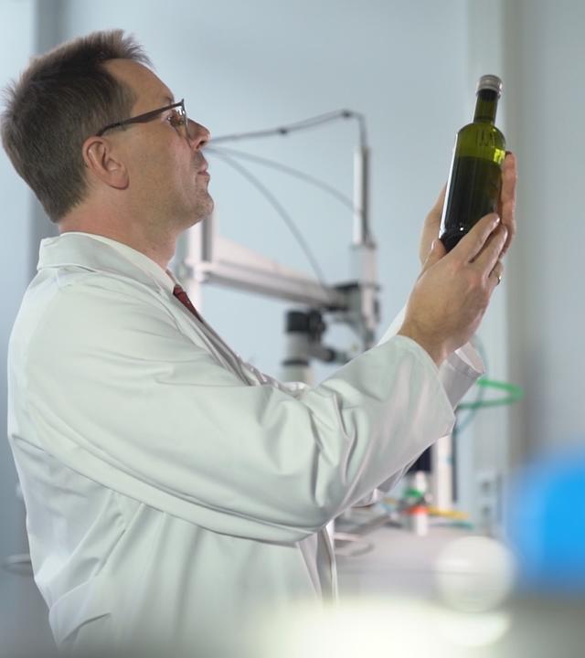 "Fake Food - Die Tricks der Lebensmittelfälscher": Prof. Stephan Schwarzinger von der Universität Bayreuth steht in einem Labor und betrachtet eine Flasche Olivenöl, die er in der Hand hält.