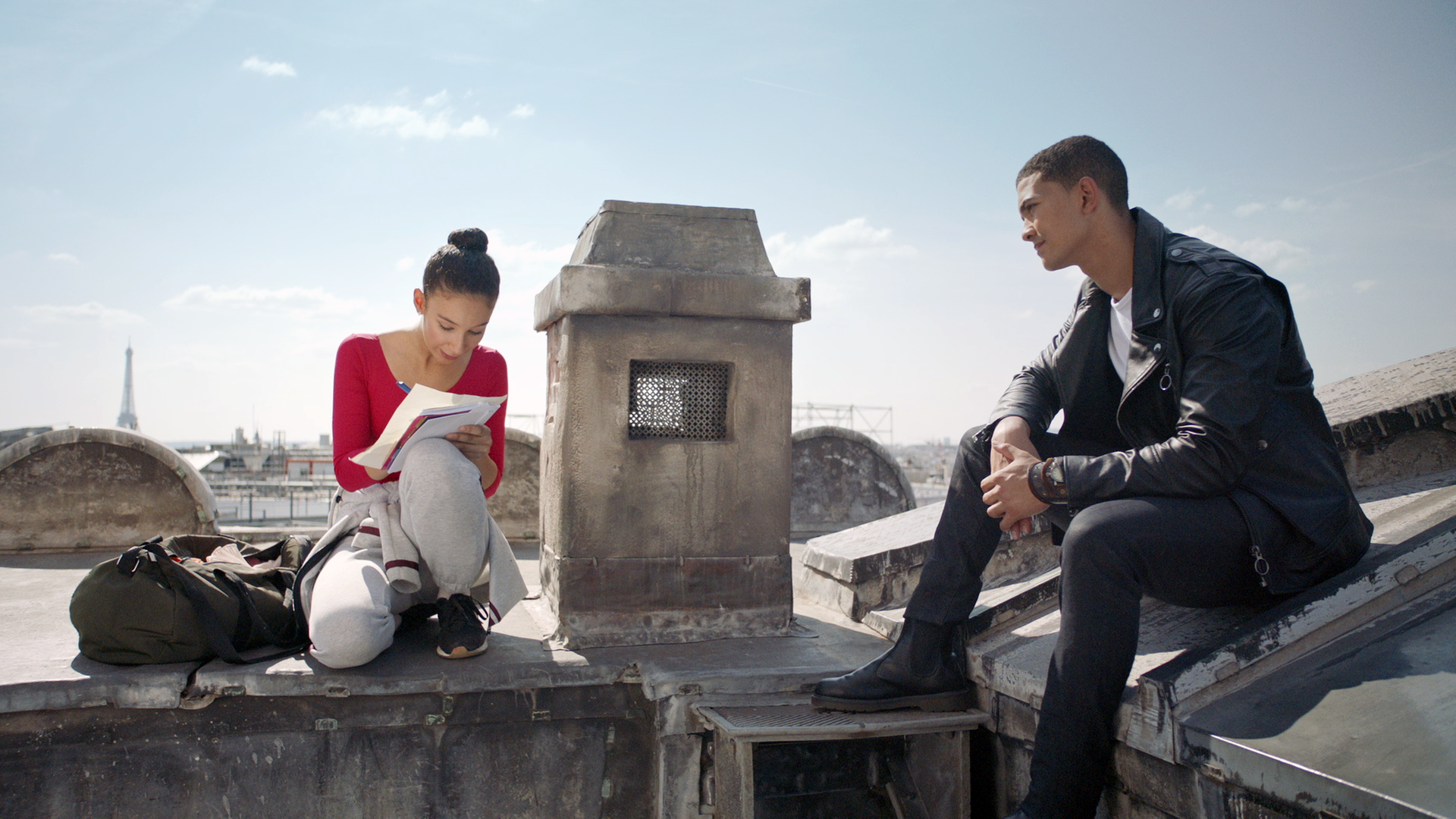 Ines (links) und Pinky (rechts) sitzen auf dem Dach der Pariser Oper. Ines schreibt etwas auf ein Blatt Paper, während Pinky ihr zusieht.
