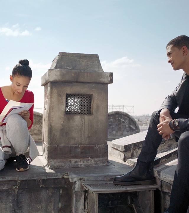 Ines (links) und Pinky (rechts) sitzen auf dem Dach der Pariser Oper. Ines schreibt etwas auf ein Blatt Paper, während Pinky ihr zusieht.