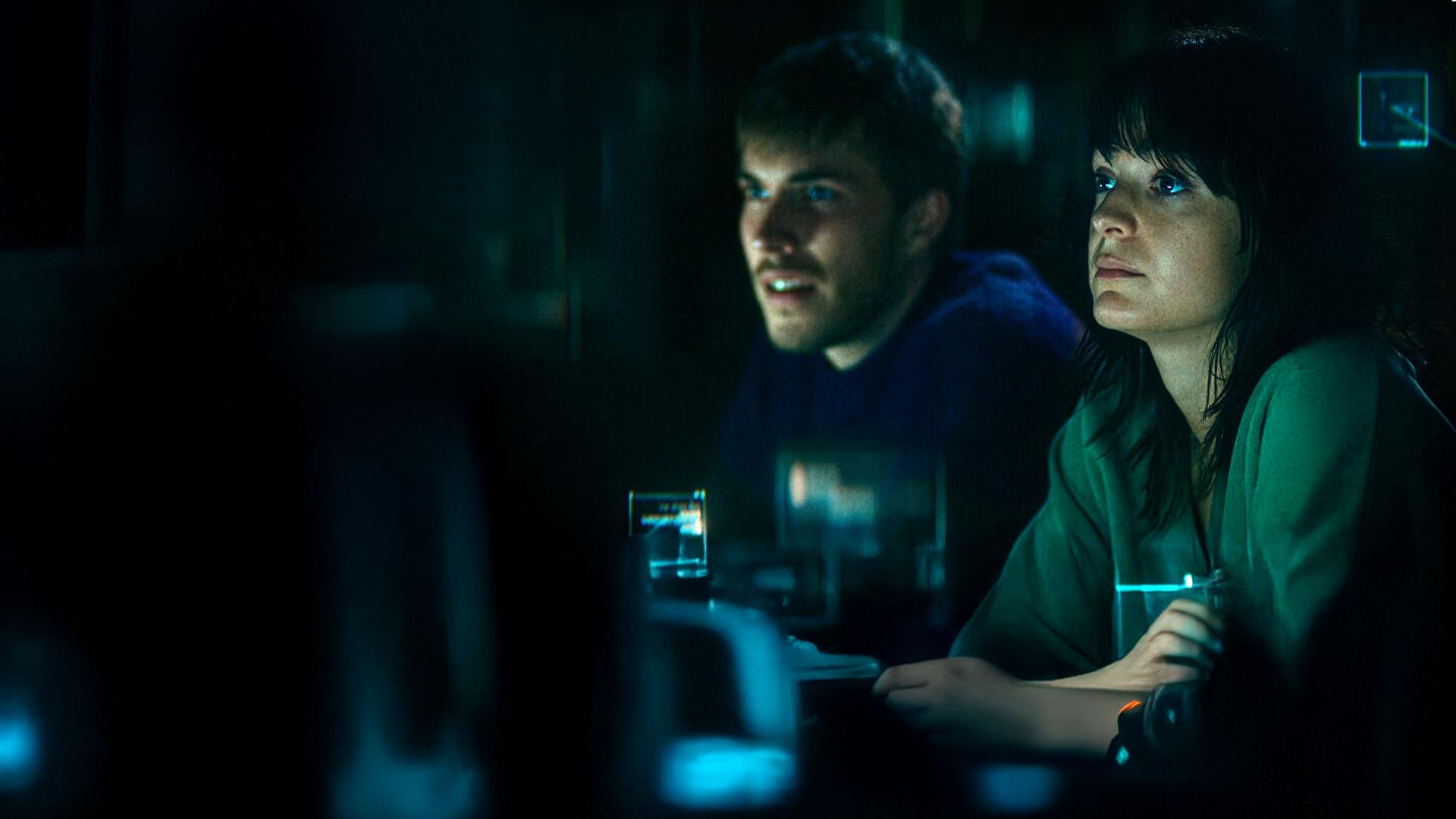 "The Kollective - Falsche Freunde": Aaron (Felix Mayr) und Delia (Céline Buckens) sitzen nebeneinander in einem dunklen Raum und beobachten etwas.