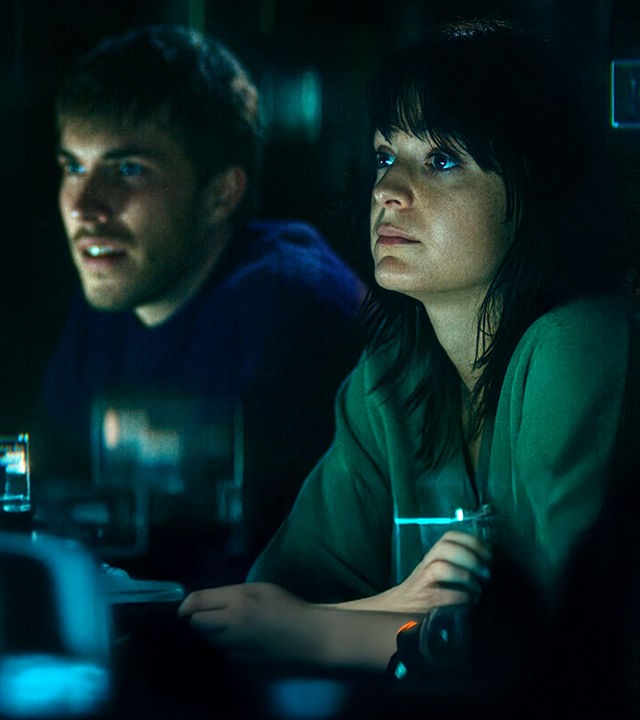 "The Kollective - Falsche Freunde": Aaron (Felix Mayr) und Delia (Céline Buckens) sitzen nebeneinander in einem dunklen Raum und beobachten etwas.