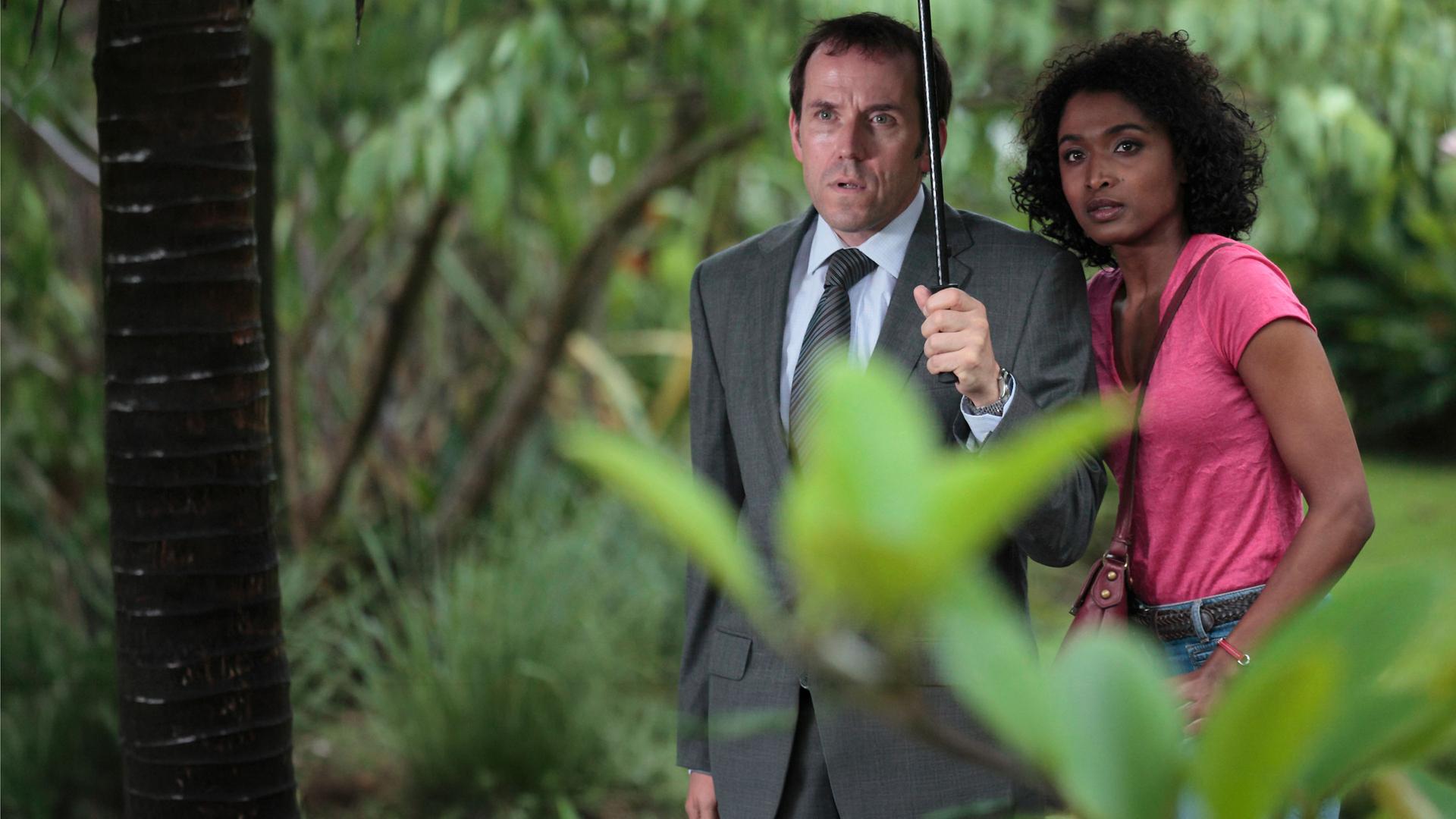 "Death in Paradise - Falsche Töne": Außenaufnahme DI Richard Poole (Ben Miller) und Camille Bordey (Sara Martins) - beide stehen unter einem Regenschirm, der von Poole gehalten wird.