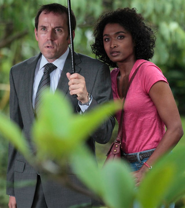 "Death in Paradise - Falsche Töne": Außenaufnahme DI Richard Poole (Ben Miller) und Camille Bordey (Sara Martins) - beide stehen unter einem Regenschirm, der von Poole gehalten wird.