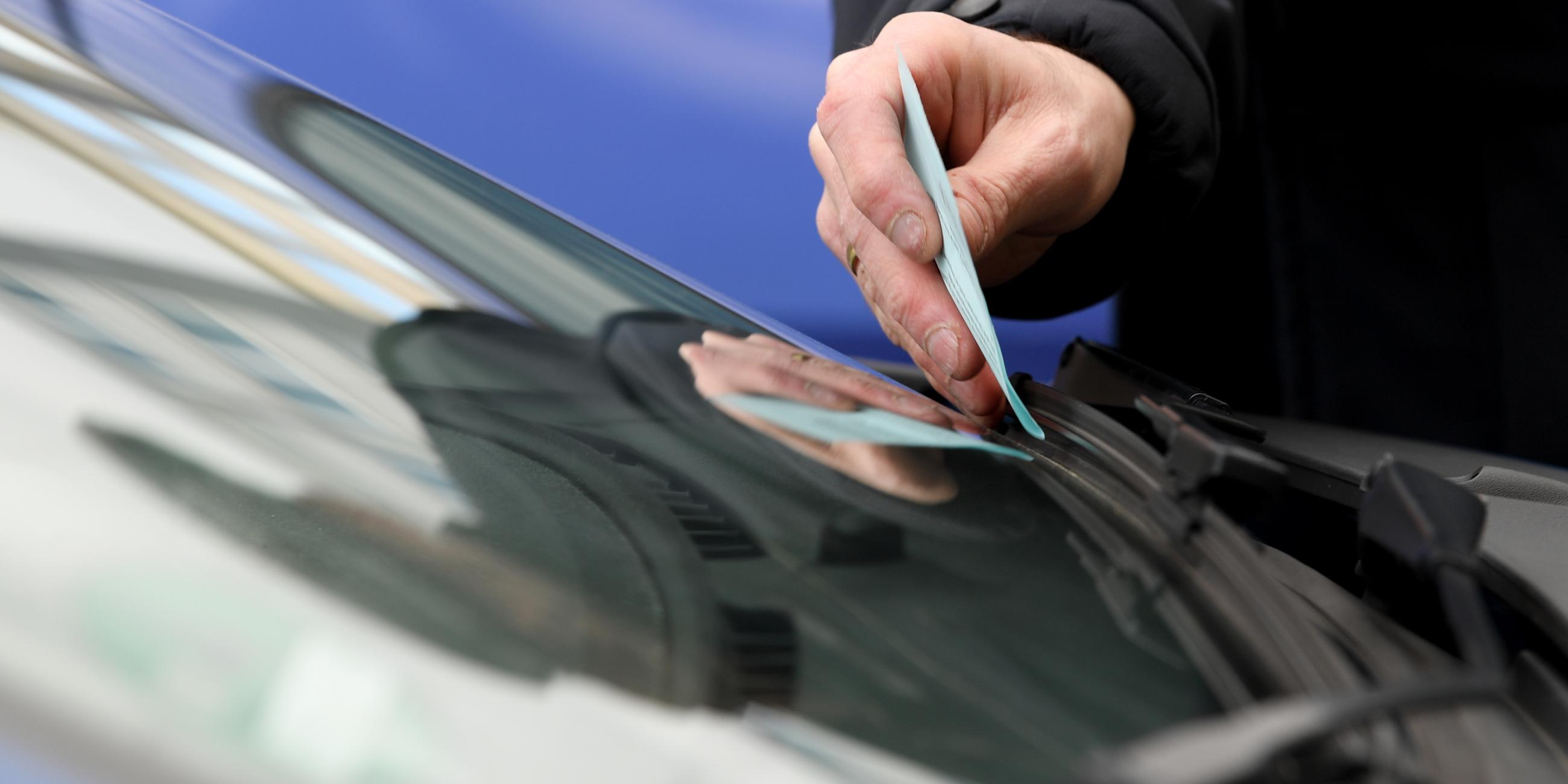 Ein Mitarbeiter der Verkehrspoilzei klemmt einen Strafzettel hinter den Scheibenwischer eines Autos.