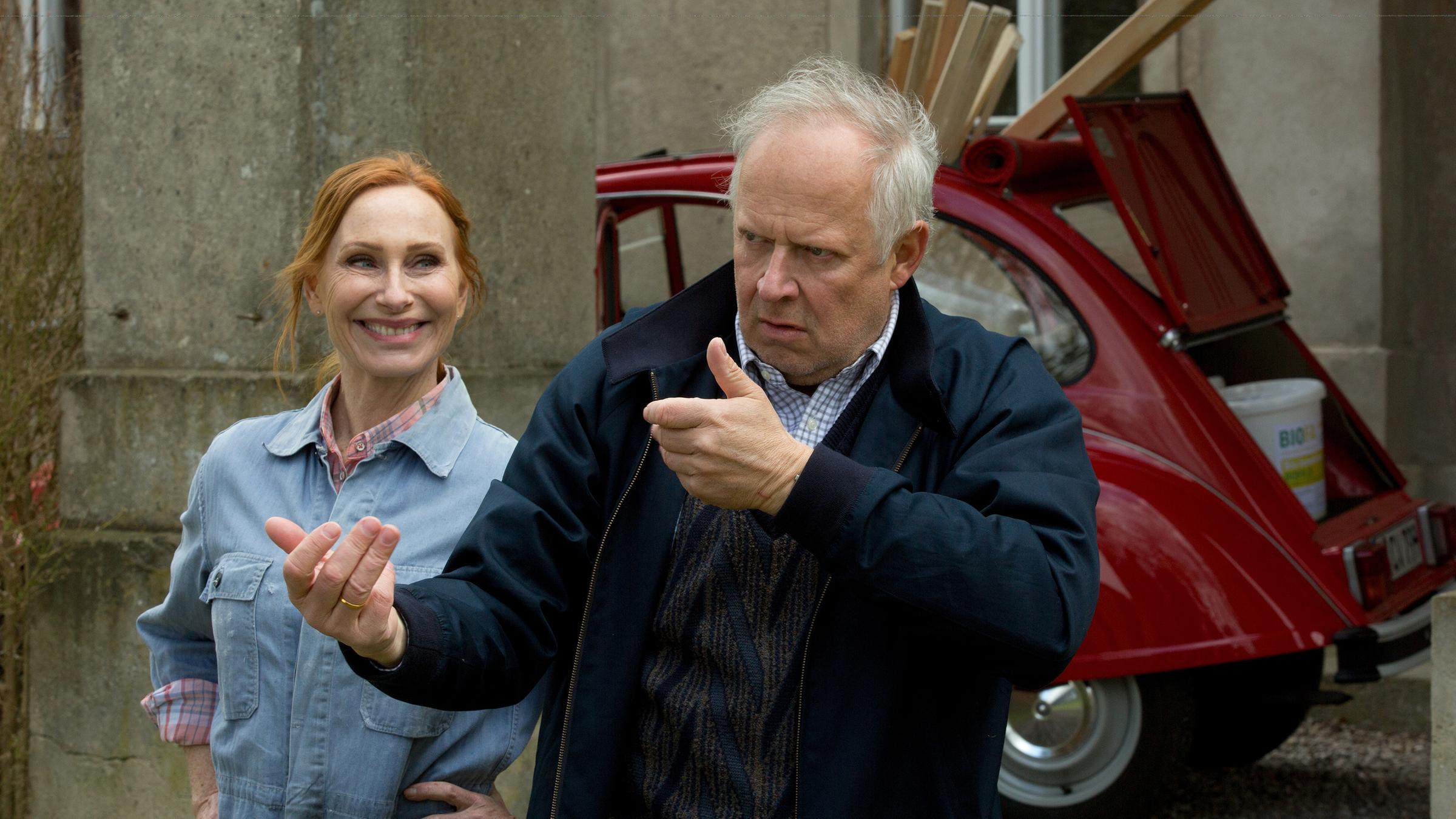 Gundula (Andrea Sawatzki, li.) himmelt ihren Gatten Gerald (Axel Milberg) an. Gerald demonstriert mit ernster Miene und großer Handgeste, wie er sich die Renovierungsarbeiten vorstellt. Beide stehen vor einer roten Ente, die mit Baumarktartikeln zugestopft ist. Im Hintergrund ist die verwitterte Hauswand des Gutshauses zu sehen.