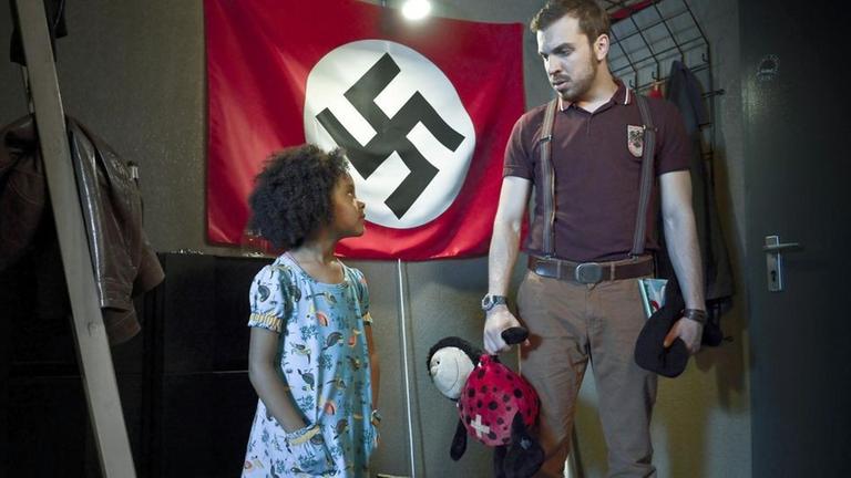 "Familie Braun": Lara (Nomie Laine Tucker) und ihr Vater Thomas (Edin Hasanovic) stehen sich im Flur der Wohnung gegenüber und schauen sich an. Thomas hält ein Buch und Laras Lieblingskuscheltier in den Händen. An der Wand hinter ihnen hängt eine Fahne mit einem Hakenkreuz.