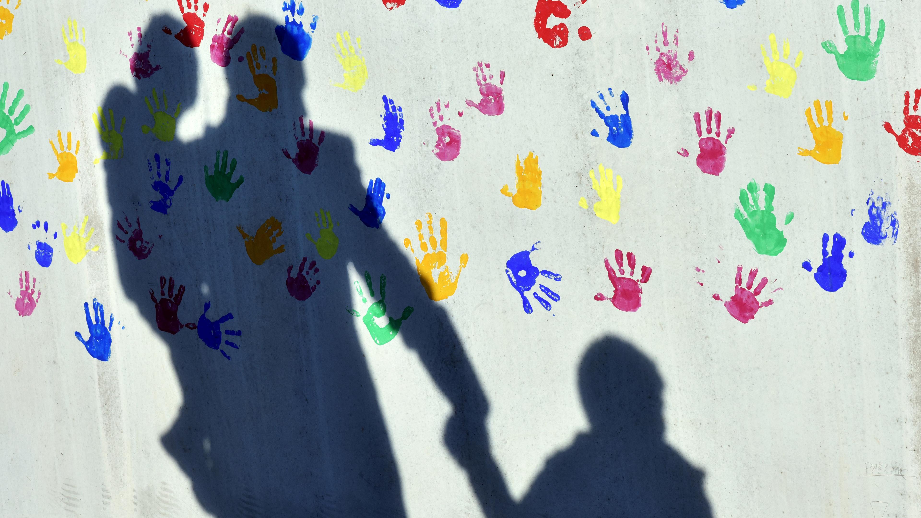 Bayern, München: Ein Mann mit einem Kind auf dem Arm und einem an der Hand wirft einen Schatten auf eine mit bunten Handabdrücken bemalte Wand einer Kindertagesstätte.