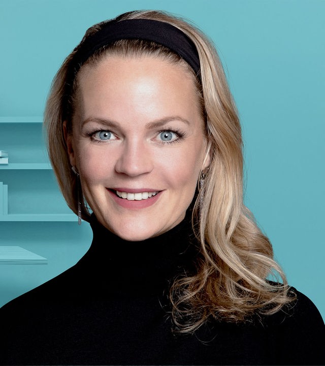 Eva Brenner im Portrait vor einem pastelblauen Hintegrund, in dem ein Büro angedeutet ist.