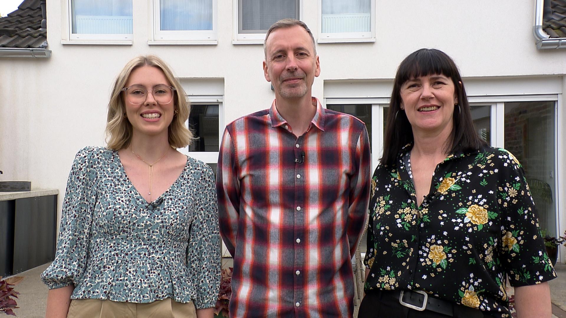 Familie Robertz, bestehend aus Vater, Mutter und Tochter, steht vor einer weißen Hauswand und lächelt in die Kamera.