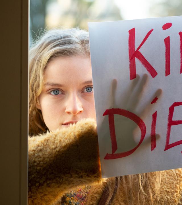 "Letzte Spur Berlin - Familienbande": Nicole Machnik (Lara Feith) steht draußen vor einem Fenster. Sie hält ein großes weißes Blatt Papier mit der Aufschrift "Kindsdiebe!" an die Glasscheibe und schaut provozierend hindurch.