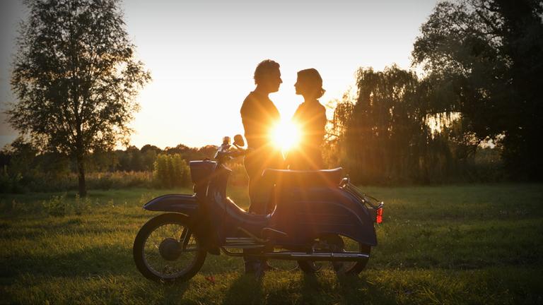 "Nächste Ausfahrt Glück - Familienbesuch": Juri Hoffmann (Dirk Borchardt) und Katharina Wegener (Valerie Niehaus) stehen im Gegenlicht eines sommerlichen Sonnenuntergangs, hinter ihnen Bäume und eine Wiese. Vor ihnen steht ein Moped der DDR-Marke "Schwalbe".