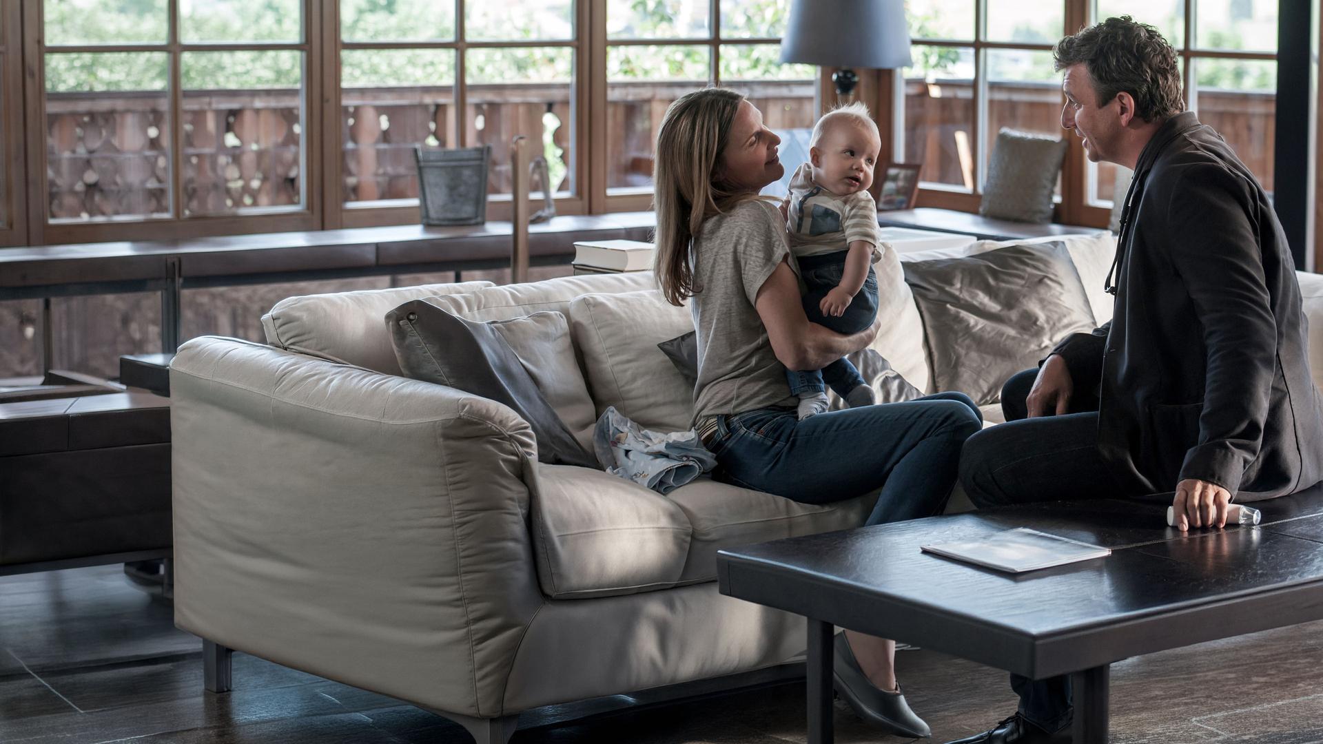 "Der Bergdoktor - Familienfieber (1)": Sabine Wimmer (Nele Mueller-Stöfen) sitzt auf der Couch und hält ihren Sohn Leo auf dem Arm. Vor ihr sitzt Dr. Martin Gruber (Hans Sigl) und betrachtet das Kind.