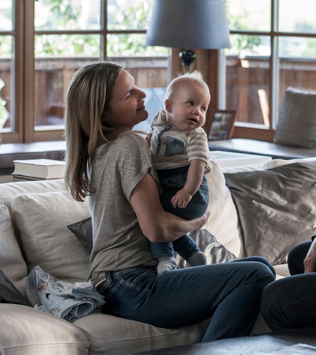 "Der Bergdoktor - Familienfieber (1)": Sabine Wimmer (Nele Mueller-Stöfen) sitzt auf der Couch und hält ihren Sohn Leo auf dem Arm. Vor ihr sitzt Dr. Martin Gruber (Hans Sigl) und betrachtet das Kind.