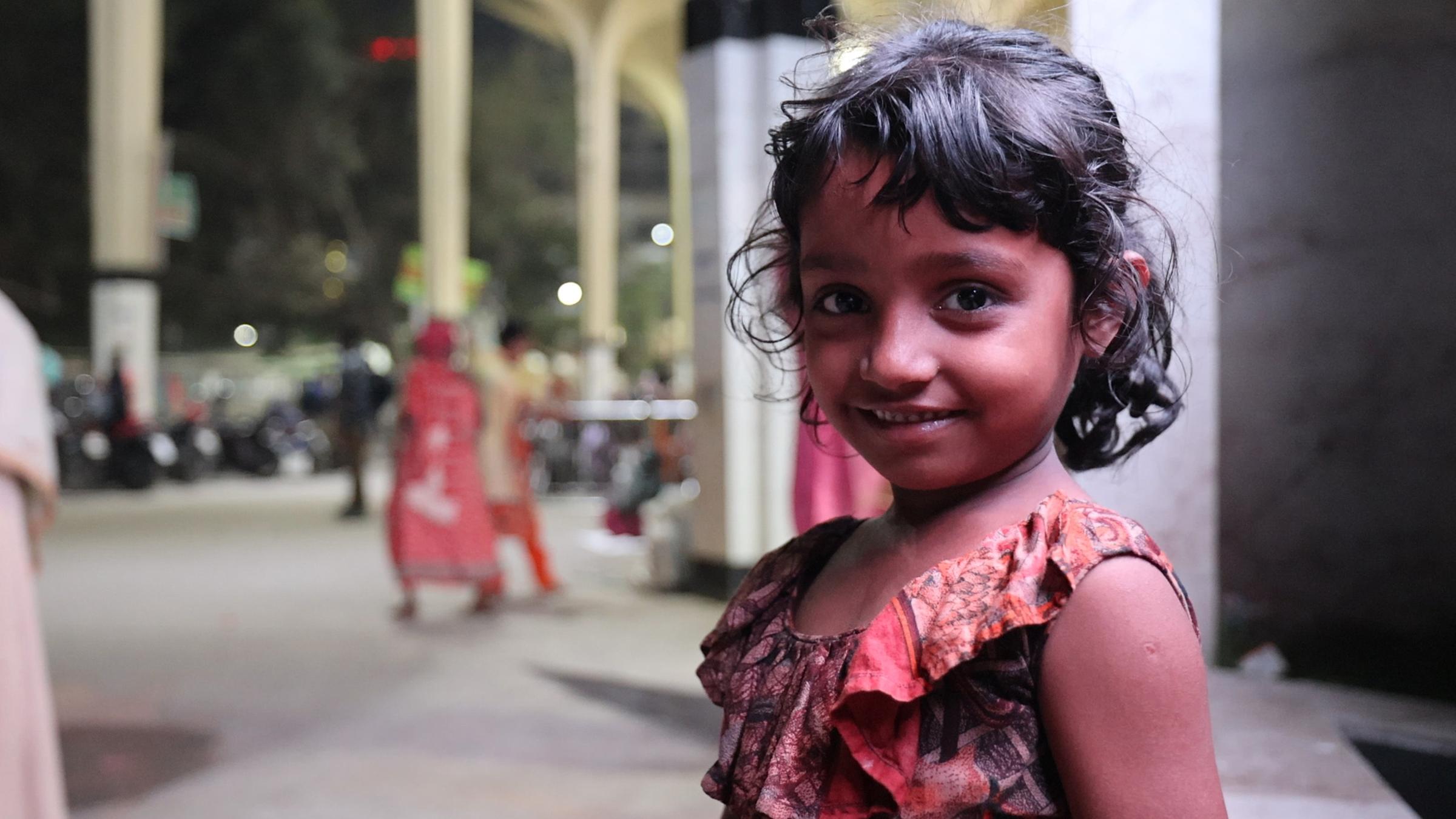 Ein kleines Mädchen mit dunklen Haaren lächelt in die Kamera. Sie trägt ein rotes Kleid mit Muster. Im Hintergrund sieht man Säulen und Menschen.