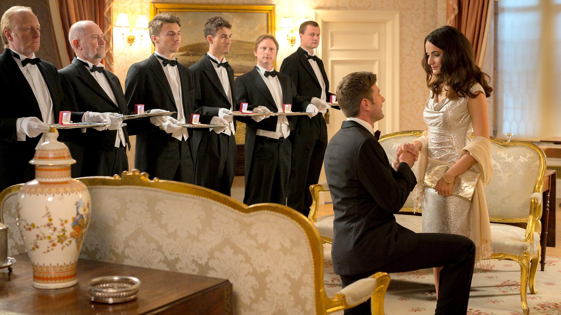 "Rosamunde Pilcher: Fast noch verheiratet": Oscar (Jochen Schropp), in schwarzem Anzug mit Fliege gekleidet, kniet vor Sienna (Paula Schramm), die ein champagnerfarbenes Abendkleid trägt, und hält ihre Hand. Beide blicken sich verliebt an. In dem nobel eingerichteten Zimmer stehen sechs Butler und halten jeweils ein Tablett, mit einer geöffneten Ringschatulle darauf, in den Händen.