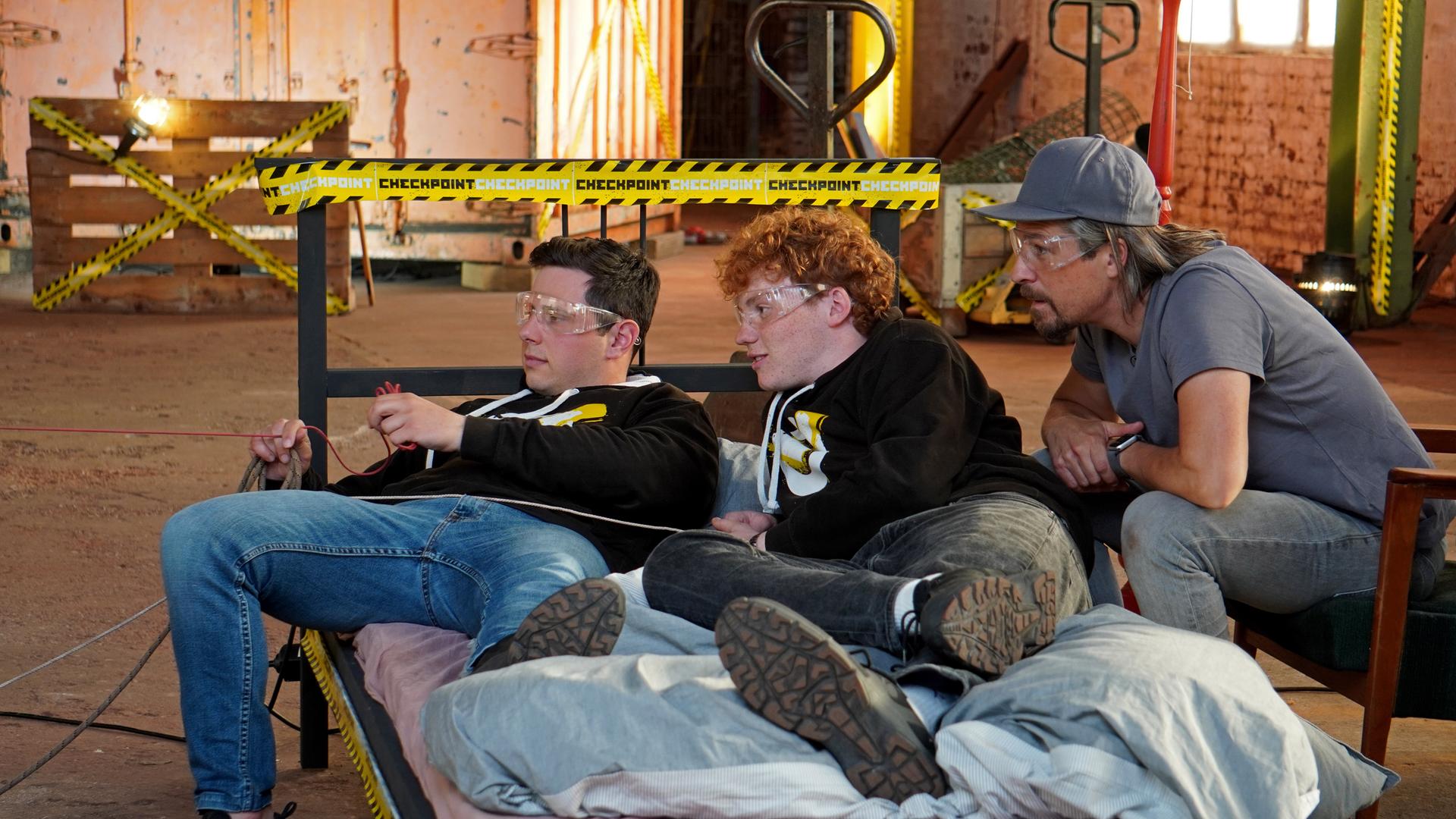 Checkpoint: Cem und Jona liegen auf dem Bett und bedienen eine Vorrichtung, die Ihnen Essen ans Bett liefern soll. Tommy Scheel schaut gebannt zu. 