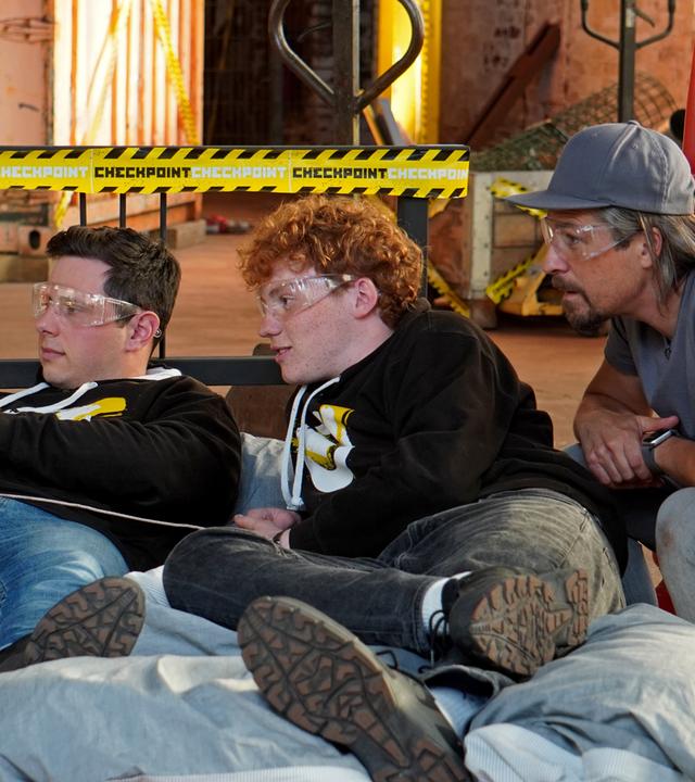 Checkpoint: Cem und Jona liegen auf dem Bett und bedienen eine Vorrichtung, die Ihnen Essen ans Bett liefern soll. Tommy Scheel schaut gebannt zu. 