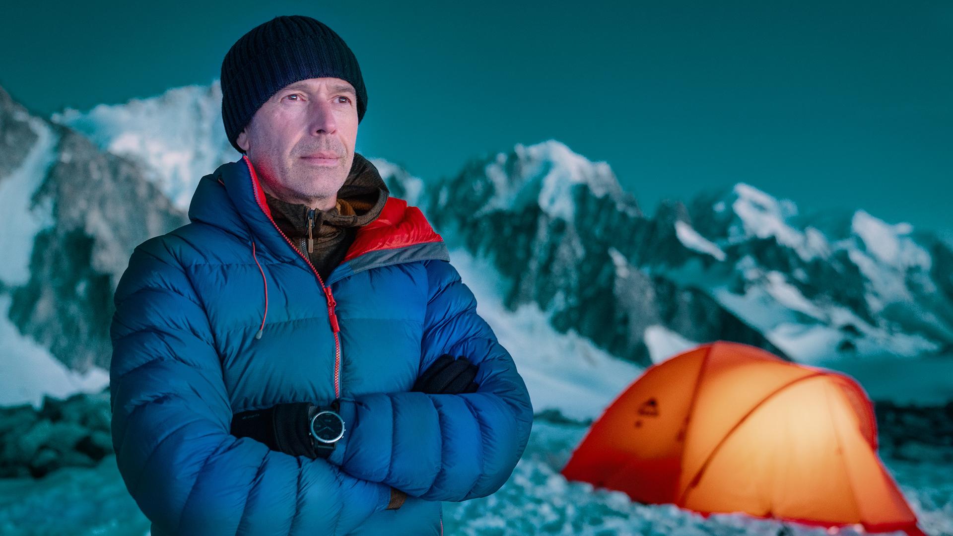 Dirk Steffens steht im Abendlicht auf dem Mont Blanc Massiv vor einem von innen heraus erleuchteten Zelt.