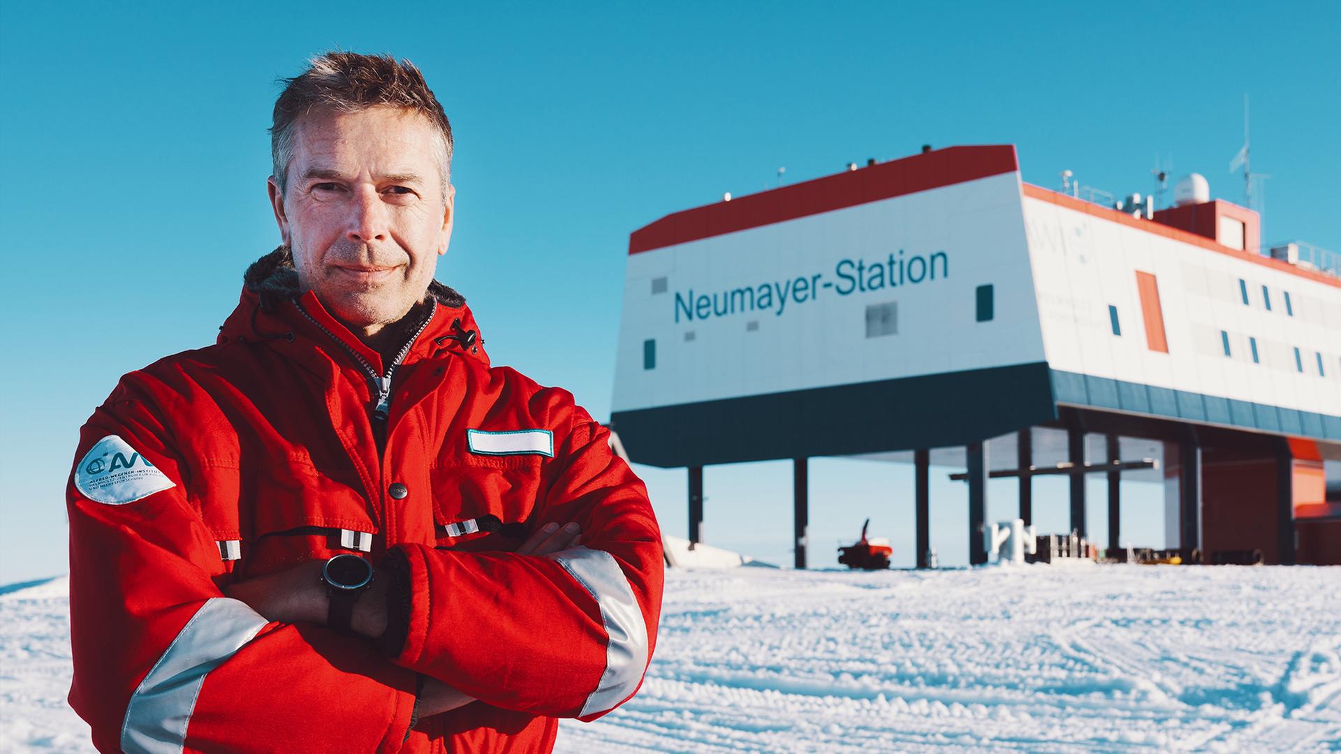 Dirk Steffens, links, steht in Schneelandschaft. Im Hintergrund, rechts, ist sie Neumayer-Station zu sehen.