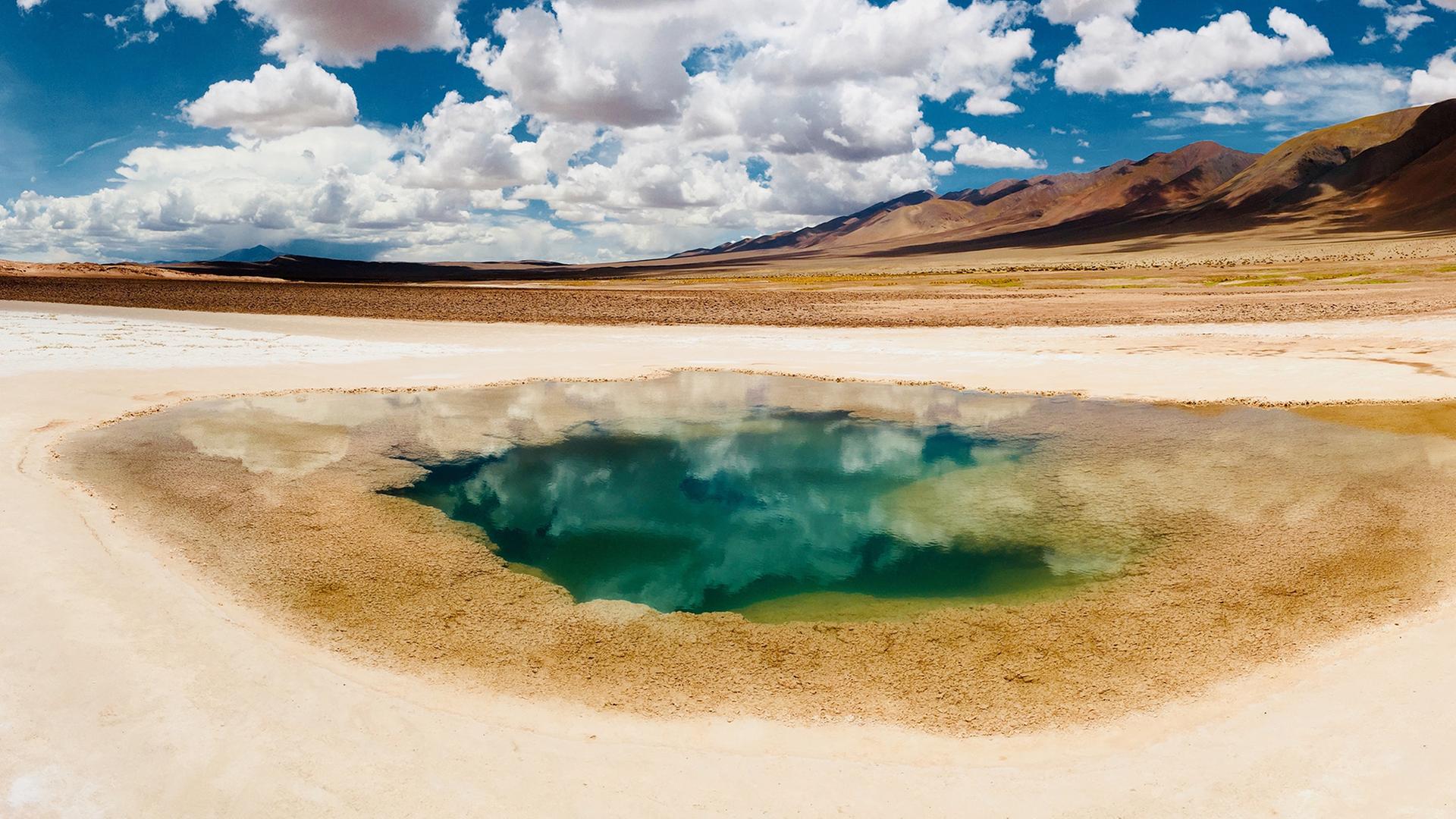 Bergige Wüstenlandschaft. Vorne mittig: Wasserkrater, in dem sich der Himmel spiegelt