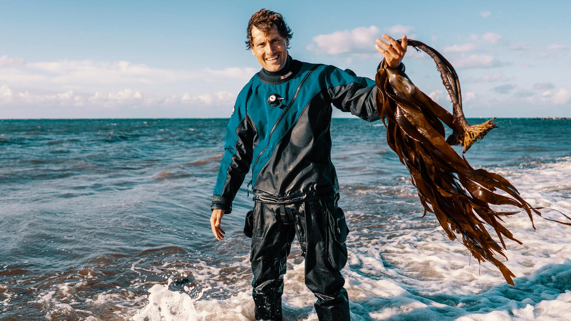 Uli Kunz im Taucheranzug steht bis zu den Knöcheln im Wasser an einem Strand. In seiner linken Hand hält er Kelp, Braunalgen.