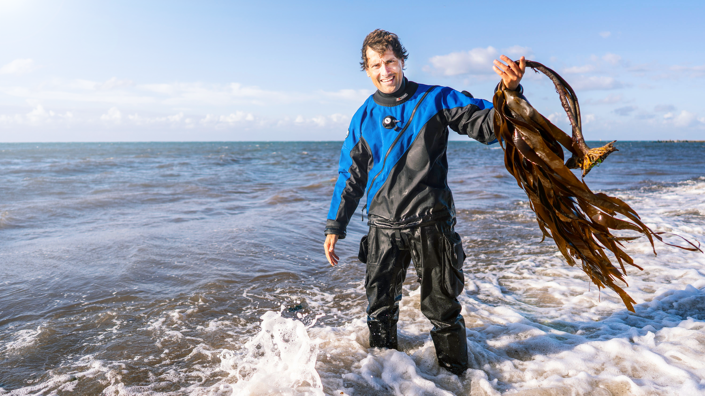 Uli Kunz im Taucheranzug steht bis zu den Knöcheln im Wasser an einem Strand. In seiner linken Hand hält er Kelp, Braunalgen.