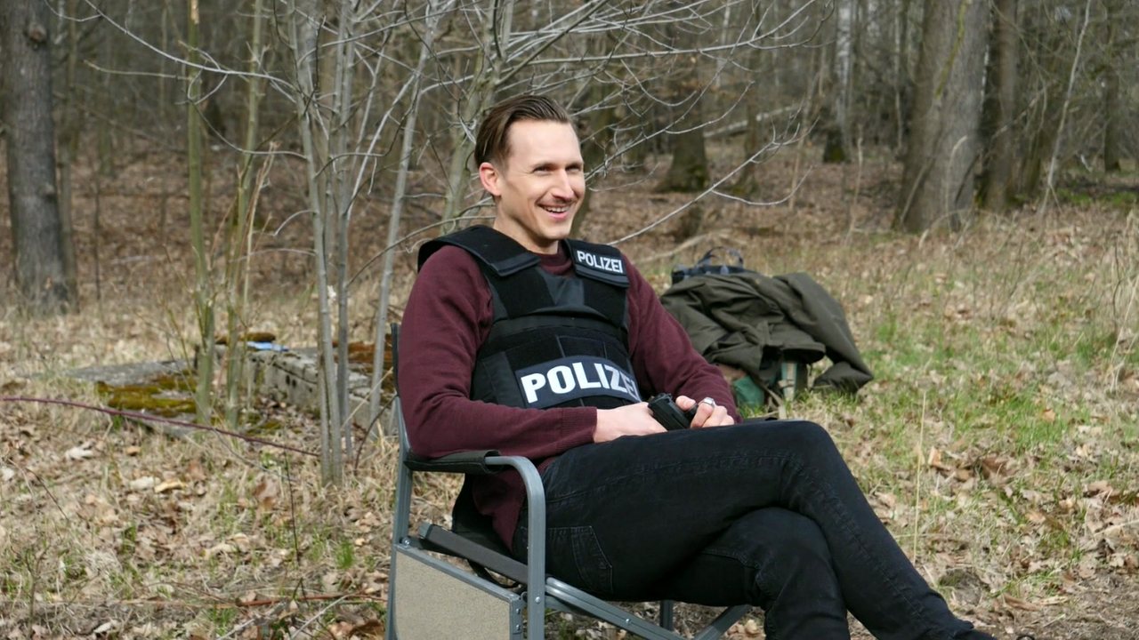 Johannes Hendrik Langer sitzt in einer Schusssicheren Polizeiweste am Set in einem Wald und lacht. In der Hand hält er eine Pistole.