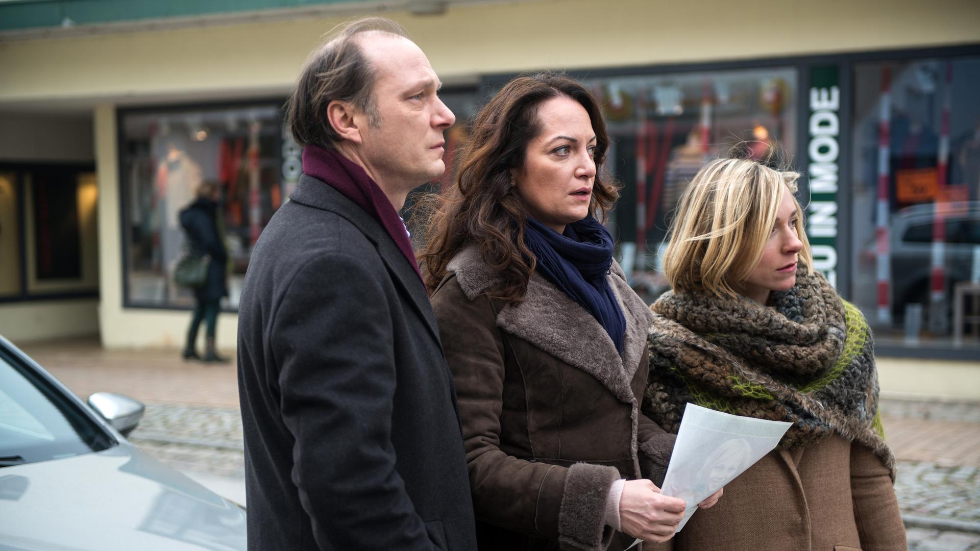 Jana (Natalia Wörner, M.), Brauner (Martin Brambach, l.) und Frida (Frederike Linke, r.) stehen auf der Straße und beobachten die Passanten. Jana hält ein Bild des vermissten Jungen in der Hand.