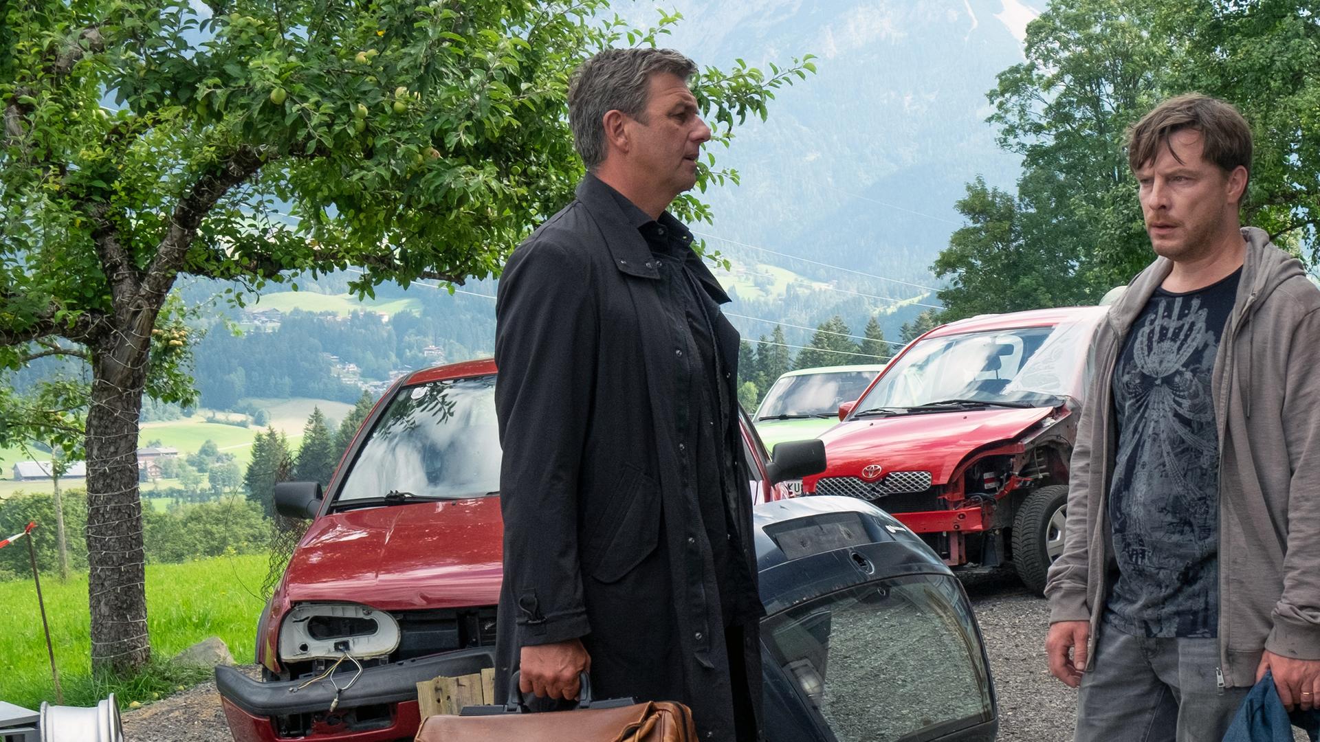 Vor einer Werkstatt stehen sich Martin (Hans Sigl) und Holger Brandstetter (Florian Panzner) gegenüber. Im Hintergrund stehen viele reparaturbedürftige Autos.