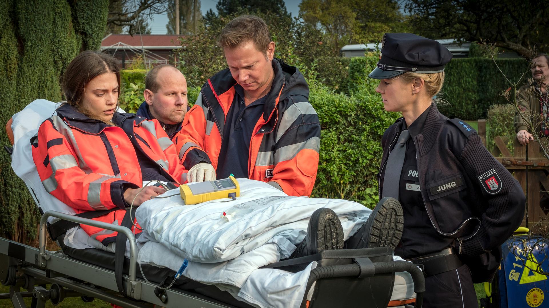 "Notruf Hafenkante - Feind und Helfer": In einem Kleingarten steht Dr. Haase (Fabian Harloff) neben einer Patiententrage, auf der Dr. Grünberg sitzt. Dr. Grünberg schaut abwesend. Am Fußende der Patiententrage steht die Polizistin Franzi Jung (Rhea Harder-Vennewald). Am Kopfende neben Dr. Haase steht ein Rettungssanitäter (Komparse).
