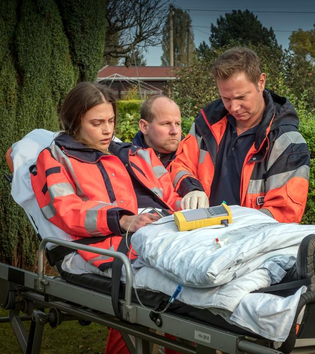 "Notruf Hafenkante - Feind und Helfer": In einem Kleingarten steht Dr. Haase (Fabian Harloff) neben einer Patiententrage, auf der Dr. Grünberg sitzt. Dr. Grünberg schaut abwesend. Am Fußende der Patiententrage steht die Polizistin Franzi Jung (Rhea Harder-Vennewald). Am Kopfende neben Dr. Haase steht ein Rettungssanitäter (Komparse).