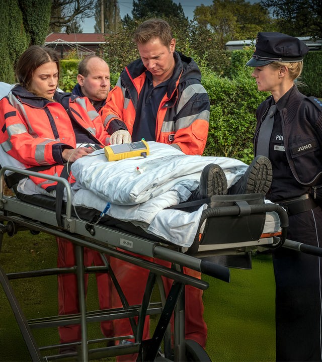 "Notruf Hafenkante - Feind und Helfer": In einem Kleingarten steht Dr. Haase (Fabian Harloff) neben einer Patiententrage, auf der Dr. Grünberg sitzt. Dr. Grünberg schaut abwesend. Am Fußende der Patiententrage steht die Polizistin Franzi Jung (Rhea Harder-Vennewald). Am Kopfende neben Dr. Haase steht ein Rettungssanitäter (Komparse).