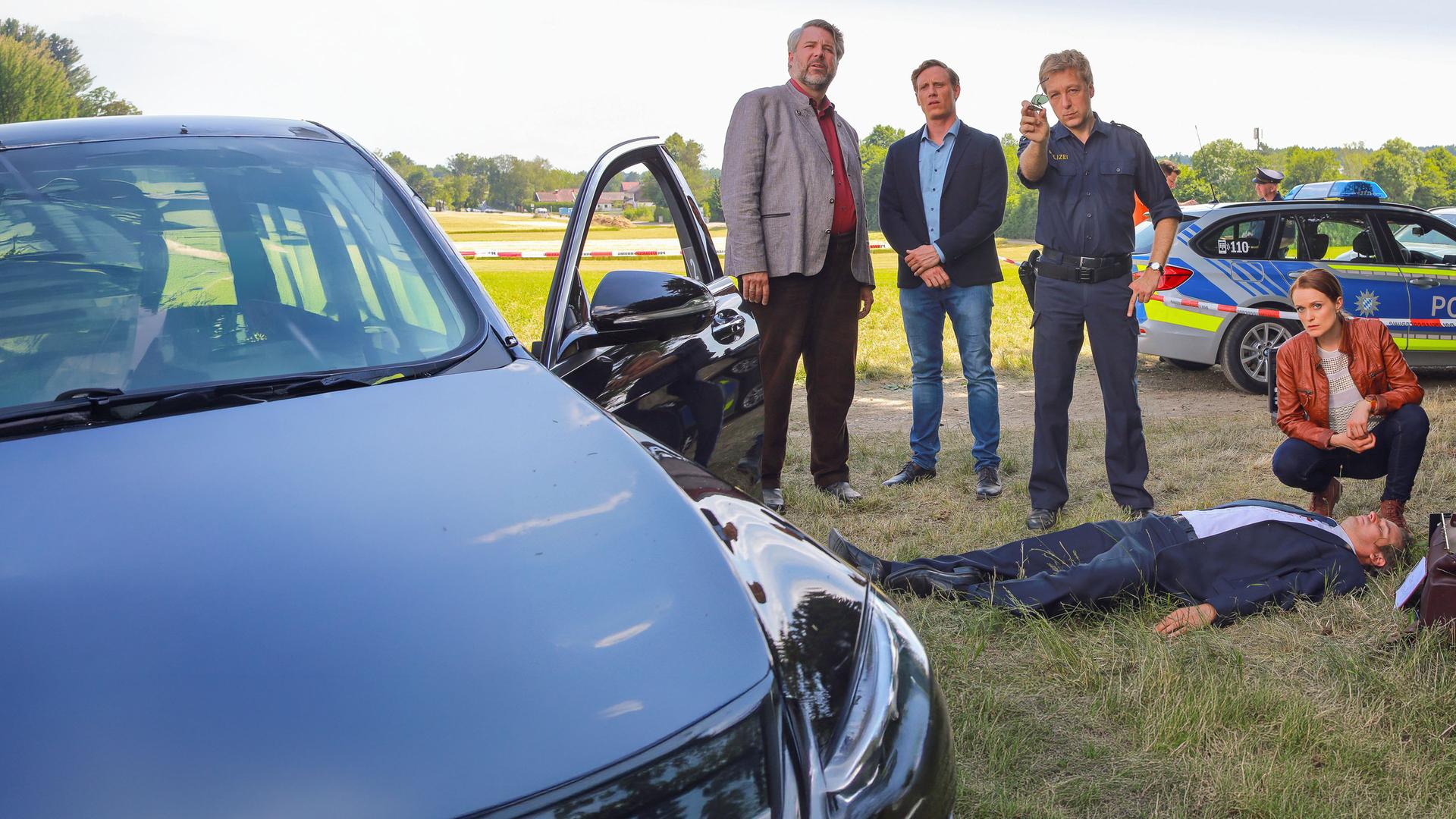 "Die Rosenheim-Cops - Feldmanns letzte Fahrt": Anton Stadler (Dieter Fischer), Christian Bach (Patrick Kalupa) und Michael Mohr (Max Müller) stehen nebeneinander am Tatort, neben ihnen hockt Sandra Mai (Sina Wilke). Vor ihnen liegt das Opfer auf dem Rücken. Sie stehen auf einer Wiese. Um sie herum sind ein Fahrzeug mit offener Fahrertür zu sehen sowie ein Polizeifahrzeug.