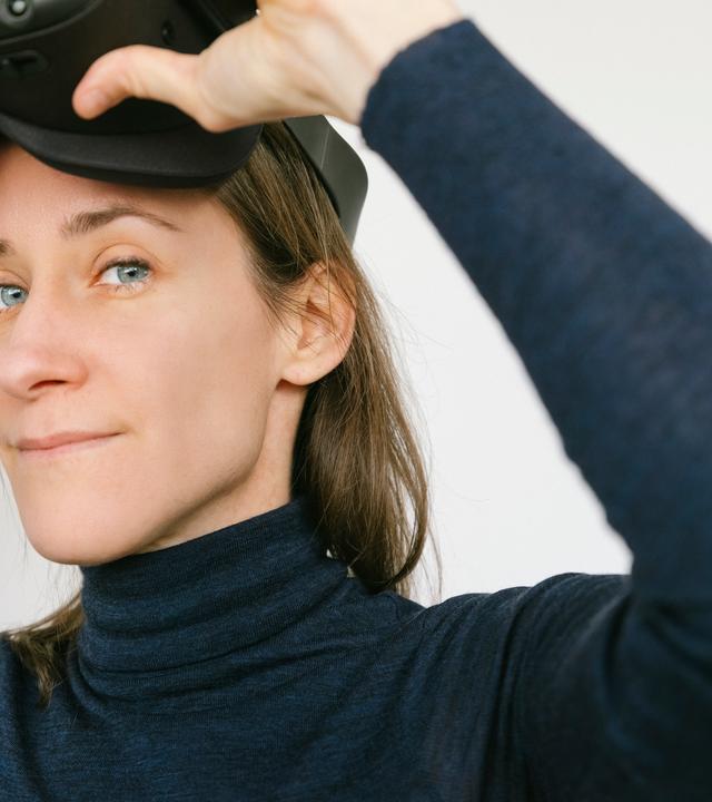 Das Bild zeigt eine Frau mit mittellangen, braunen Haaren. Sie trägt ein enges, dunkles Oberteil mit einem hohen Kragen. Ihr Blick ist konzentriert, während sie ein VR-Headset in beiden Händen hält und dabei ist, es auf ihren Kopf zu setzen. Der Hintergrund ist hell und neutral, wodurch die Aufmerksamkeit auf die Frau und das Headset gelenkt wird. Die Umgebung ist schlicht, ohne auffällige Elemente. Es handelt sich um eine Momentaufnahme, die den Einsatz von Virtual-Reality-Technologie suggeriert.