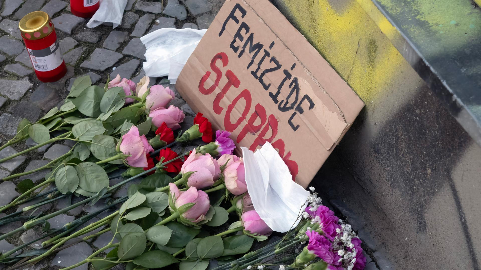 Ein Pappschild mit der Aufschrift "Femizide stoppen" steht angelehnt auf Kopfsteinpflaster. Davor liegen Blumen.