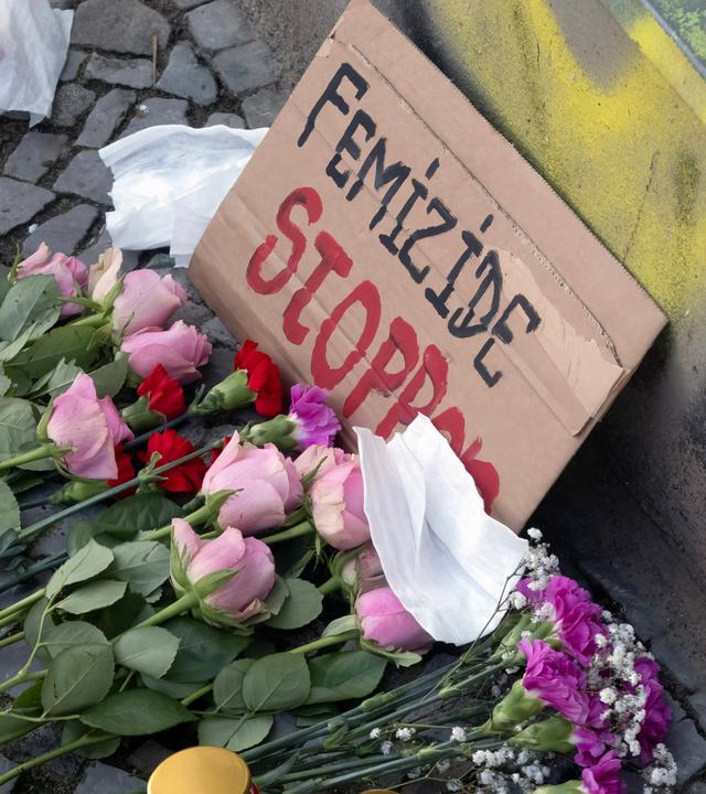 Ein Pappschild mit der Aufschrift "Femizide stoppen" steht angelehnt auf Kopfsteinpflaster. Davor liegen Blumen.