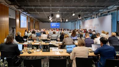 Weitwinkelaufnahme einer ZDF-Fernsehratssitzung im März 2026. Zahlreiche Menschen sitzen an langen Tischen, die zu einem offenen Rechteck angeordnet sind. Vor ihnen stehen Mikrofone, Namensschilder und Getränke. Am hinteren Ende des Saals ist eine Leinwand mit ZDF-Logos zu sehen