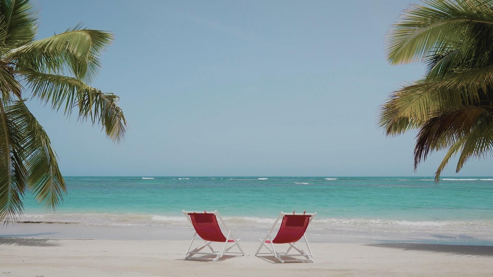  Zu sehen sind zwei rote Liegestühle am Strand. Im Hintergrund das Meer und links und rechts Palmen.