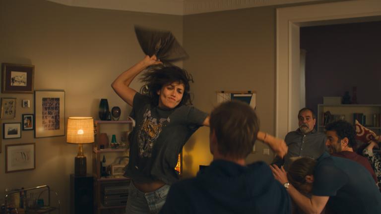 Karo (Lucie Heinze) steht in Wohnzimmer und holt mir einem Kissen in der Hand in Richtung Knut (Helgi Schmid) aus, der mit dem Rücken zur Kamera vor ihr sitzt. Karsten (Richard Kreutz), Bernhard (Matthias Brenner), Hannes (Mohamed Achour) sitzen daneben und schauen erschrocken zu.