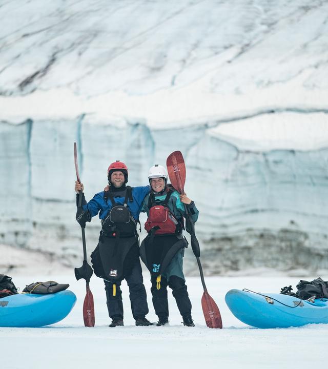 Zwei Personen mit Paddeln in der Hand stehen zwischen Kanus in eisiger Landschaft.