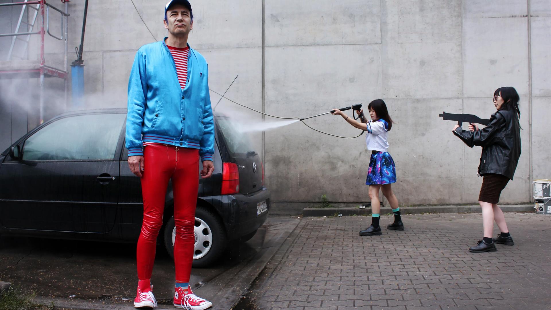 Ein Mann in roter Hose und mit einer blauen Jacke bekleidet, steht vor einem Auto. Rechts daneben steht eine Frau, die das Auto wäscht und eine Frau, die eine Maschinenpistole auf das Auto richtet.