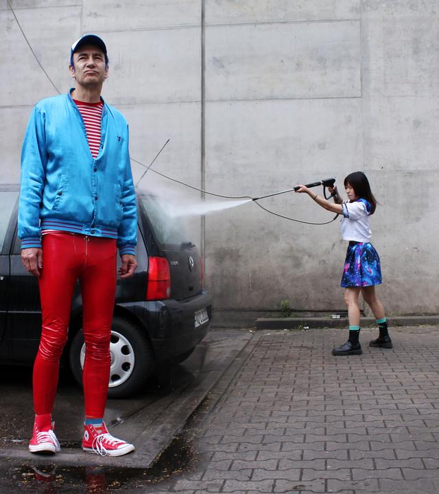 Ein Mann in roter Hose und mit einer blauen Jacke bekleidet, steht vor einem Auto. Rechts daneben steht eine Frau, die das Auto wäscht und eine Frau, die eine Maschinenpistole auf das Auto richtet.