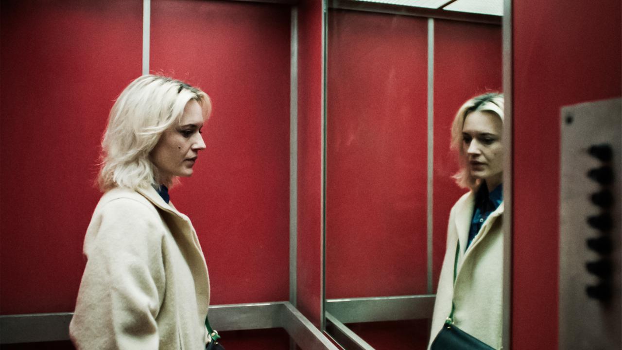 Man sieht eine Frau in einem Fahrstuhl, die sich in einem großen Spiegel betrachtet.