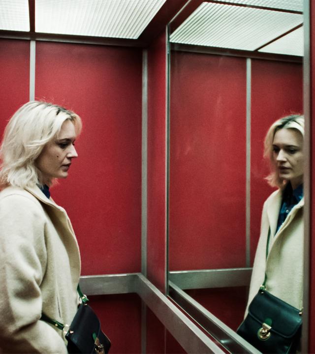 Man sieht eine Frau in einem Fahrstuhl, die sich in einem großen Spiegel betrachtet.
